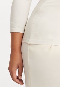 Krämfärgad topp med trekvartslång ärm, med rundad nederkant och subtil logotypdetalj på sidan. Mjuk och slät tygtextur.