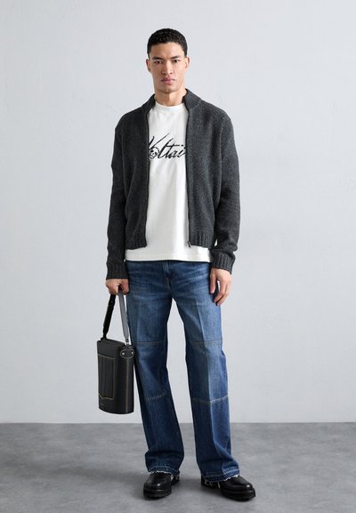 Un gilet en tricot zippé gris porté sur un t-shirt graphique blanc, associé à un pantalon en denim bleu et des chaussures à plateforme noires, tenant un sac noir.