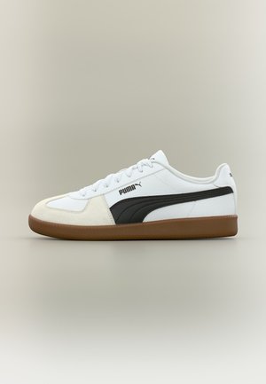 Biely nízky teniska Puma s čiernym pruhom, béžovou semišovou špičkou, bielymi šnúrkami a hnedou gumovou podrážkou, zobrazený z boku na jednoduchom pozadí.