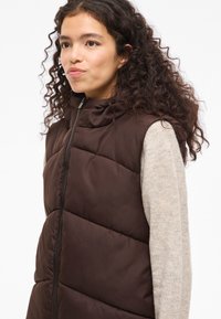 Smeđa podstavljena prsluk s visokim ovratnikom, s prednjim zatvaračem na patentni zatvarač i bešavnim dijagonalnim šavovima. Nošen preko svijetlo bež teksturiranog džempera.