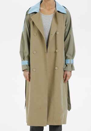 Persoon die een lange beige trenchcoat draagt met een lichtblauwe kraag en mouwbanden, over een grijs geribd shirt en donkere broek.