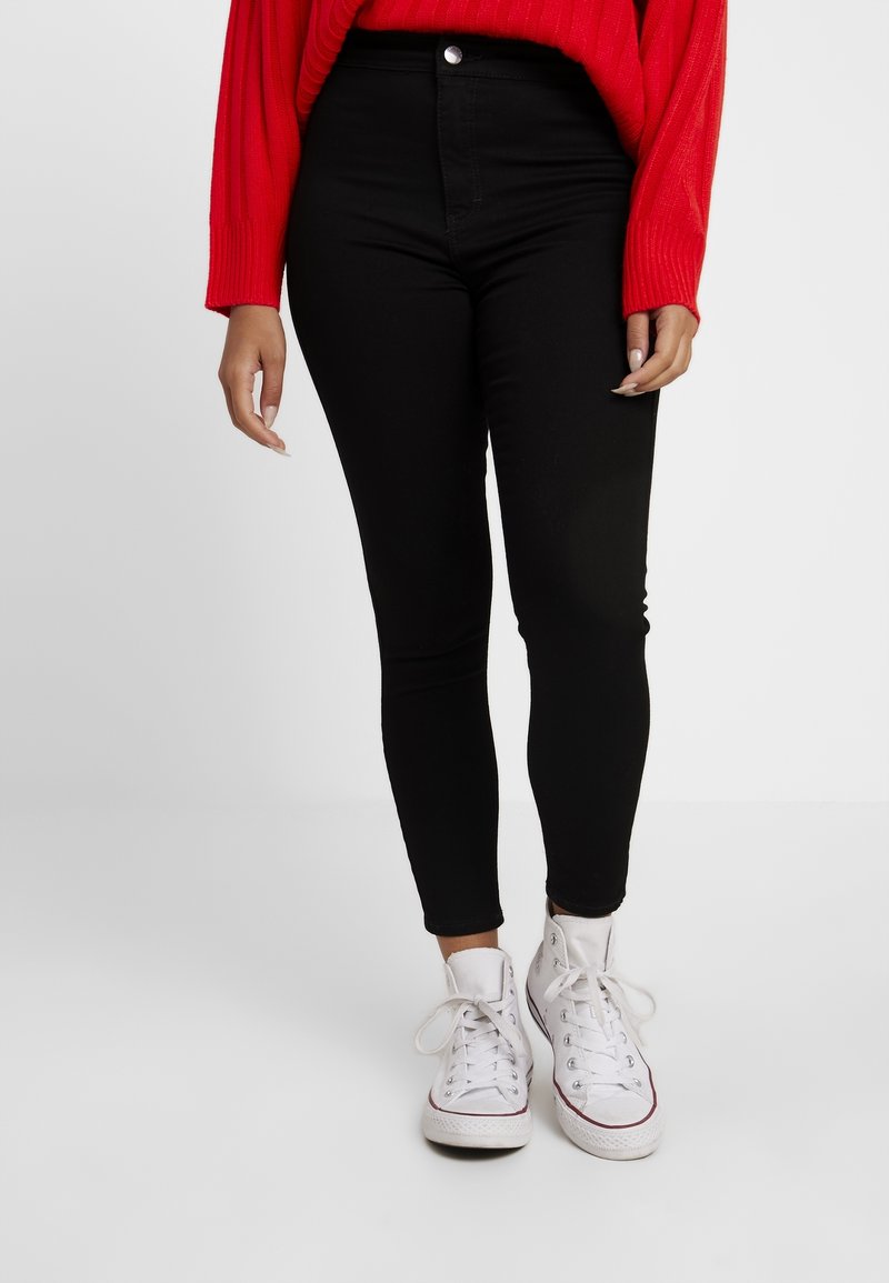 Jeans negros de cintura alta con un ajuste slim, ligero elasticidad, cierre frontal con botón y diseño de cinco bolsillos, combinados con zapatillas blancas.