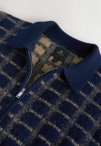 Modrý a béžový károvaný sveter na zips vyrobený z mäkkej vlnenej zmesi, s golierom a lesklým strieborným zipsom. Viditeľná značka značky vo vnútri.