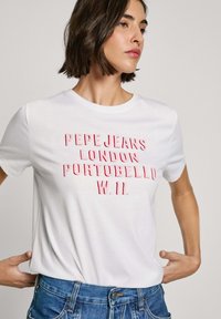 Balta medvilnė marškinėliai su ryškiu raudonu iškilmingu tekstu: "PEPE JEANS LONDON PORTOBELLO W.11." Trumpomis rankovėmis, klasikinio apvalaus kaklo dizainas.