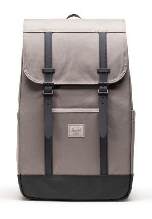 Sac à dos en tissu gris avec des straps et des boucles noires, ainsi qu'un fond noir, comportant une poche avant et une petite étiquette de marque rectangulaire.