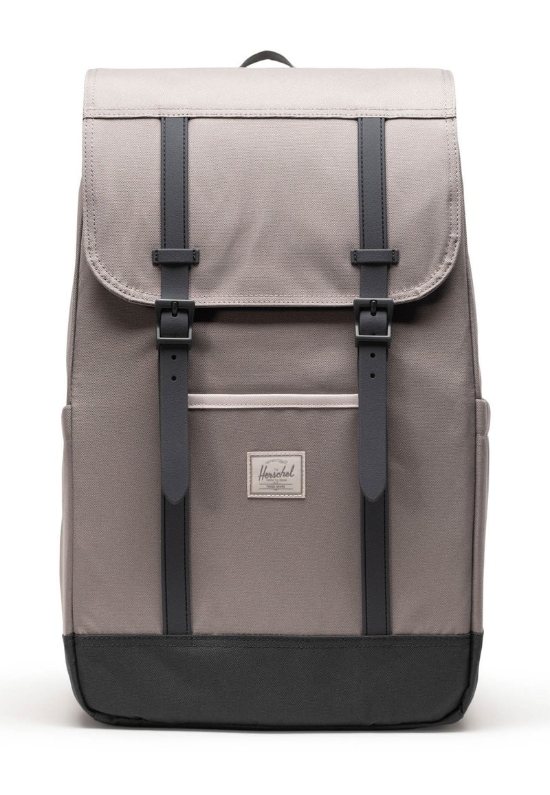 Mochila de tela gris con correas, hebillas y parte inferior negras, que cuenta con un bolsillo frontal y una pequeña etiqueta de marca rectangular.