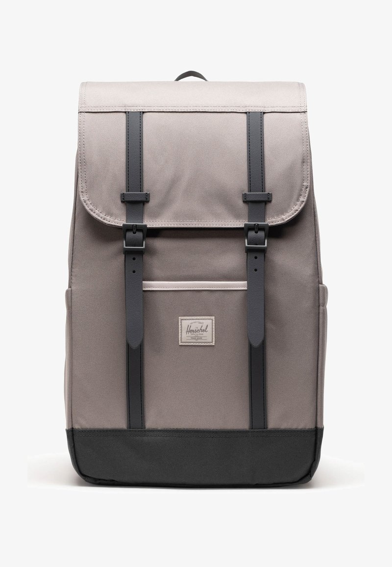 Mochila de tela gris con correas, hebillas y parte inferior negras, que cuenta con un bolsillo frontal y una pequeña etiqueta de marca rectangular.