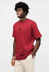 Μπορντό βαμβακερό t-shirt με στρογγυλή λαιμόκοψη και κοντά μανίκια, με μικρό μαύρο λογότυπο Nike στο στήθος. Συνδυάζεται με μπεζ παντελόνι.