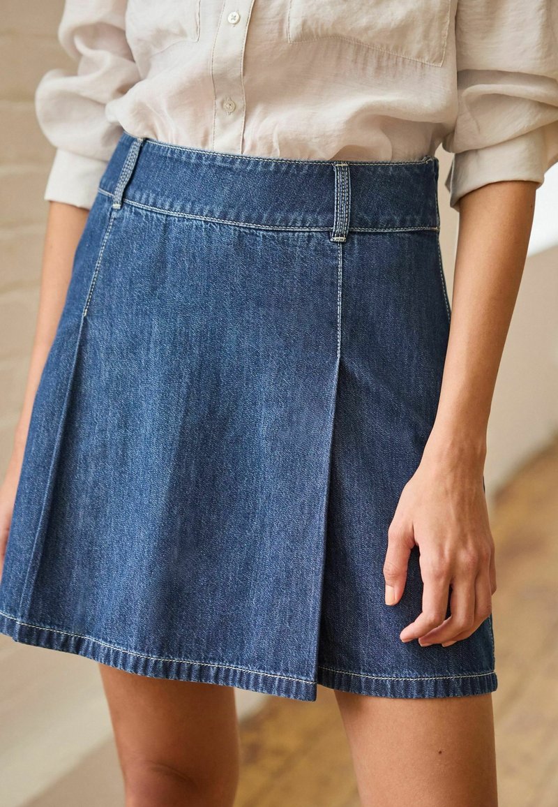 Denimvetari, jossa on A-linjainen muotoilu, raaka helma ja etukaitale. Varustettu vyötärönauhalla, jossa on vyölenkit; keskisininen väri vaalennettulla tekstuurilla.