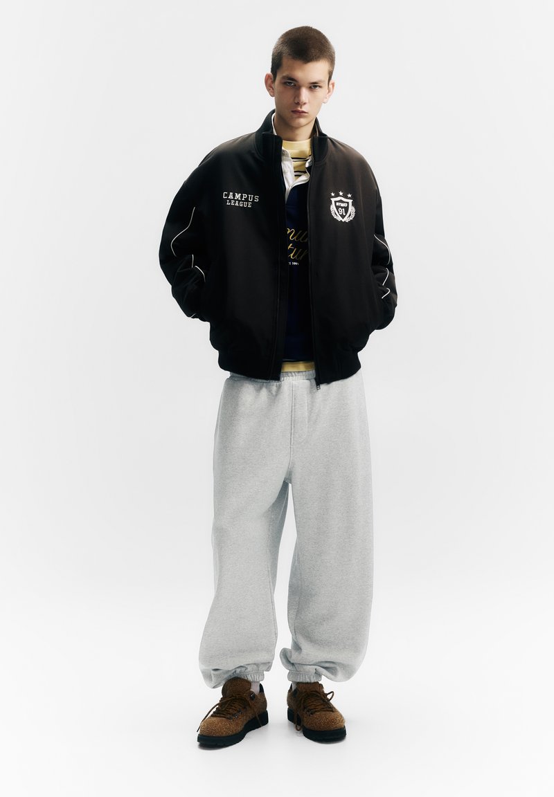 Jachetă bomber neagră cu accente albe, pantaloni de trening gri și adidași maro cu finisaj texturat. Detalii ale cămășii suprapuse sunt vizibile.