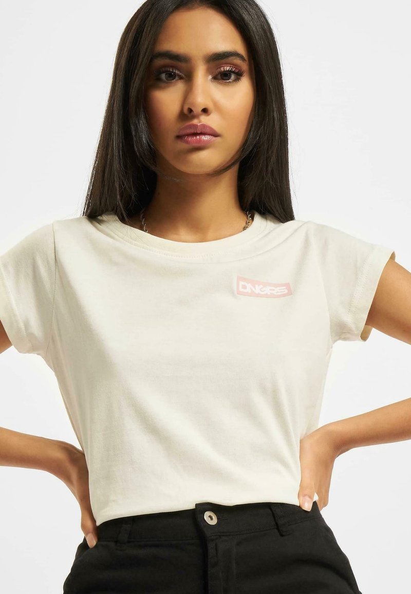 Tricou din bumbac de color creme, cu mâneci scurte, prezentând o patch cu logo roz. Asortat cu pantaloni negri cu talie înaltă pentru un look casual.