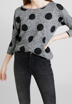 Kobieta ubrana w szary sweter z rękawami do łokci z dużymi czarnymi grochami i wysokim stanem czarnych dżinsów na jednolitym tle.