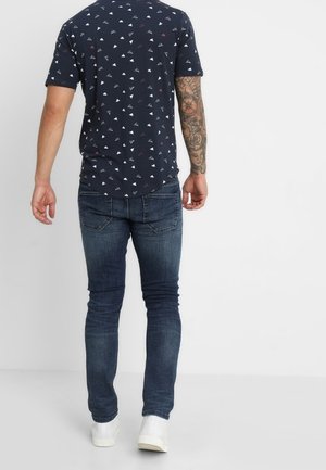 Man wearing navy patterned t-shirt, dark blue slim jeans, white sneakers, with a full-sleeve tattoo on his right arm, standing against a white background.