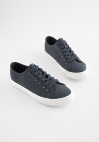 Tmavomodré plátenné tenisky s bielymi gumovými podrážkami a zodpovedajúcimi šnúrkami. Majú textúrovaný vrch a zaoblený tvar špičky.