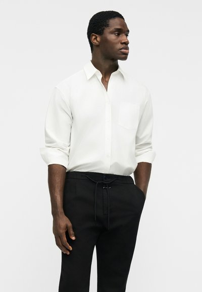 Άνδρας που φοράει λευκό πουκάμισο με μακριά μανίκια κουμπωμένο, με τυλιγμένα μανίκια και μαύρο παντελόνι με κορδόνι στη μέση, κοιτώντας προς τα πλάγια.