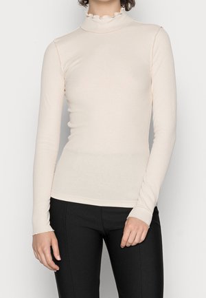 Vrouw draagt een aansluitende, crèmekleurige, langermige geribde top met een gerimpelde col en zwarte aansluitende broek.