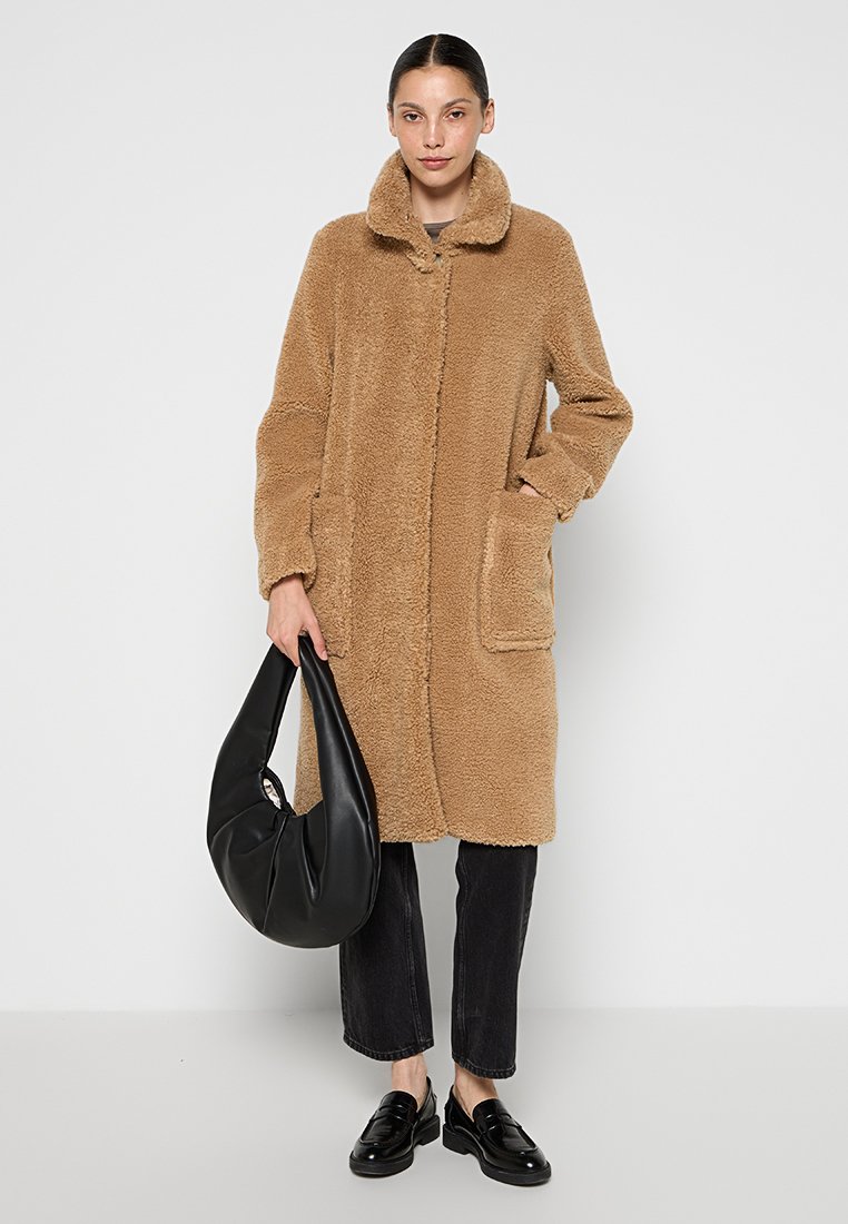 Manteau teddy beige et duveteux avec un col et des poches avant, associé à un jean noir et un sac à main noir décontracté. Mocassins lisses et brillants.