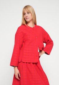 Czerwona teksturowana kurtka z okrągłym kołnierzem, zapięciem na trzy guziki i szerokimi rękawami, z subtelnym paskowanym wzorem oraz dopasowaną spódnicą.