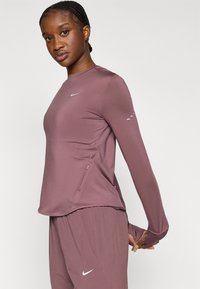 Αθλητικό τοπ με μακριά μανίκια σε χρώμα μωβ, κατασκευασμένο από λείο ύφασμα με στρογγυλή λαιμόκοψη. Χαρακτηριστικό λογότυπο Nike στο στήθος και λεπτομέρεια με φερμουάρ στο πλάι.