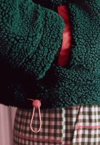 Roheline tekstuuriga fliisjope punase sisemise voodriga, millel on roosa luku tõmblukk servas, ühes ruudulise seelikuga.