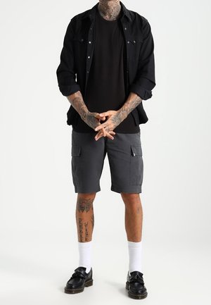 Camisa negra abotonada sobre una camiseta negra, pantalones cortos cargo grises con bolsillos, calcetines blancos, zapatos negros y tatuajes visibles en brazos y piernas.