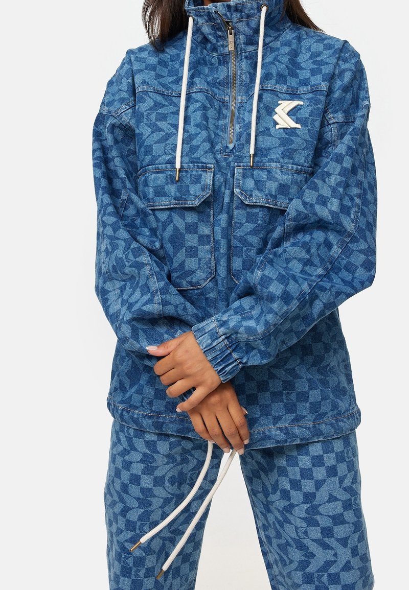 Veste en denim bleue avec un motif géométrique, col haut, fermeture éclair et deux poches avant avec des cordons blancs.