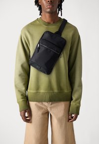 Sort crossbody taske lavet af nylon, med to lynlåslommer og en stof skulderrem, båret over en olivengrøn sweatshirt.