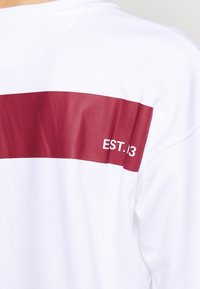 Vit t-shirt med ett horisontellt vinrött rand över ryggen där "EST.13" är tryckt i vitt. Tyget ser mjukt ut med en slät struktur.