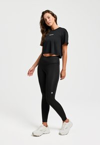 Zwarte cropped t-shirt met korte mouwen; zwarte high-waisted leggings; wit logo op leggings; gecombineerd met witte sportschoenen.