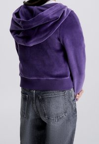 Fialová mikina s měkkou texturou, vybavená kapucí a žebrovaným lemem, spárovaná se šedými džínami s zadními kapsami.
