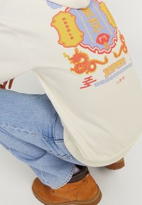 Person som bär ljusblå jeans, beige läderstövlar och en krämfärgad sweatshirt med en färgglad röd och gul drake samt ett Japan-temat grafiskt tryck.