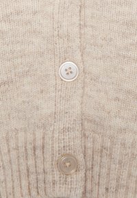 Beige gestrickte Strickjacke mit einer gerippten Textur, ausgestattet mit zwei hellen Knöpfen mit jeweils vier Löchern, die sich an der Vorderseite befinden.