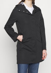 Czarny wodoodporny płaszcz z kapturem, zatrzaskami i bocznymi kieszeniami; charakteryzuje się gładką, matową teksturą i lekko błyszczącym kołnierzem.