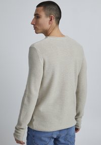 Helles beige, langärmliges, strukturiertes Strickoberteil mit rundem Halsausschnitt und seitlichen Schlitzen, kombiniert mit blauen Jeans. Dezente Nahtdetails sind sichtbar.