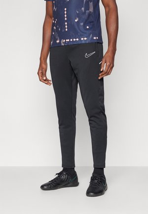 Mann trägt schwarze Nike Trainingshose, schwarze Sportschuhe und ein dunkelblaues gemustertes Hemd, steht vor einem einfarbigen hellen Hintergrund.