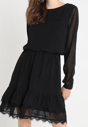 Frau trägt ein schwarzes Kleid mit transparenten langen Ärmeln und Spitzenbesatz am Saum, stehend vor einem schlichten hellen Hintergrund.