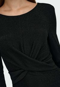 Czarna sukienka z teksturowanej, błyszczącej tkaniny. Posiada szeroki dekolt, długie rękawy oraz skręcony detal z przodu podkreślający kształt i design.