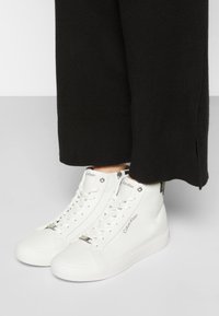 Bílé vysoké tenisky Calvin Klein z hladké kůže, se šněrováním, detailem loga a stříbrným kováním. Texturovaná gumová podrážka a aerodynamický design.