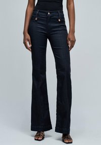 Vaqueros de tiro alto en denim negro, acampanados, con bolsillos frontales decorativos, doble cierre de botón y un acabado liso, ligeramente texturizado.
