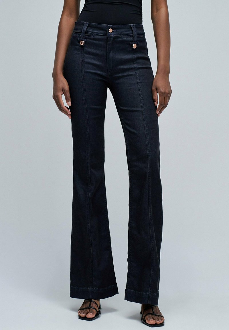 Vaqueros de tiro alto en denim negro, acampanados, con bolsillos frontales decorativos, doble cierre de botón y un acabado liso, ligeramente texturizado.