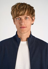 Marinblå texturerad zip-jacka med hög krage över en enkel vit t-shirt. Jackan har en synlig dragkedja och ribbad krage.