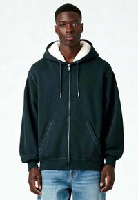 Must värviga kapuutsiga nahkjakker, millel on pehme valge vooder, kaks eesmist taskut, nööriga kapuuts ja soonikuga varrukad. Kanti siniste teksade peale.