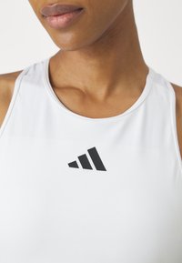 Gros plan sur une personne portant un haut sans manches blanc avec un logo noir à trois bandes centré sous l'encolure.