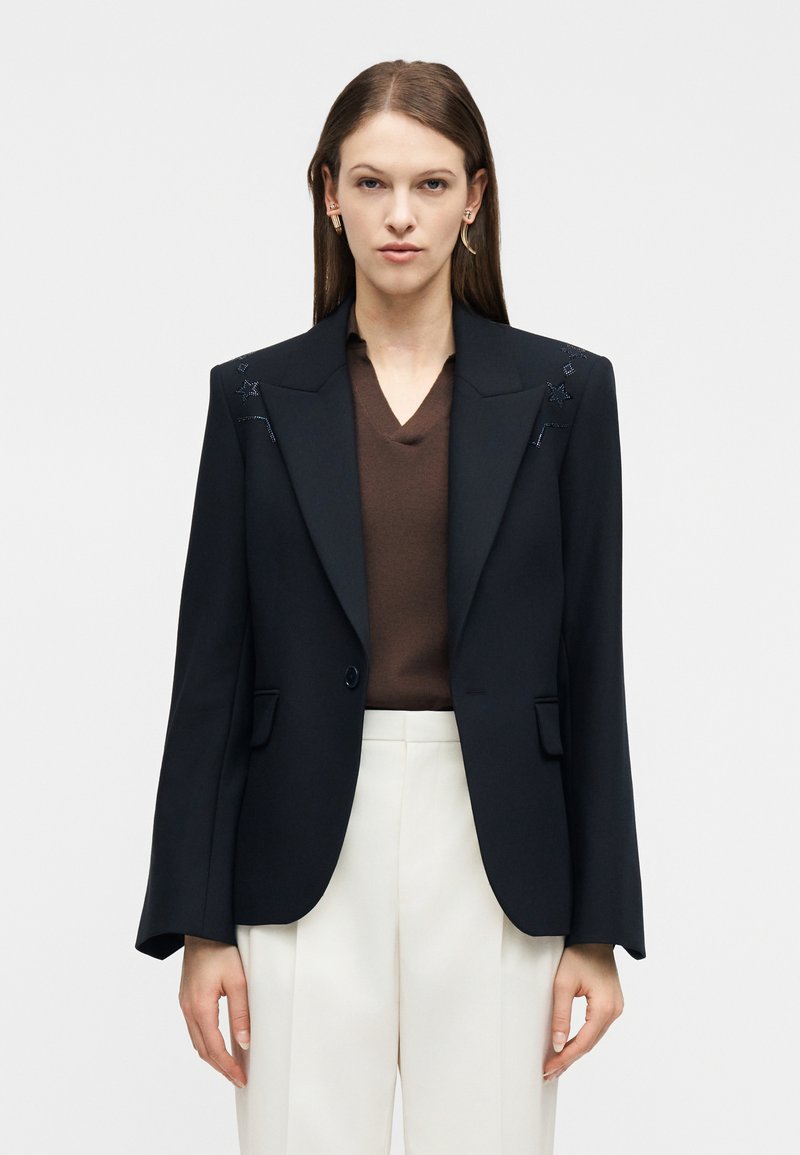 Γυναίκα που φοράει μαύρο προσαρμοσμένο σακάκι με διακοσμητικά αστεράκια, καφέ μπλούζα με V λαιμόκοψη, λευκό παντελόνι και χρυσά σκουλαρίκια, στέκεται μπροστά σε λευκό φόντο.