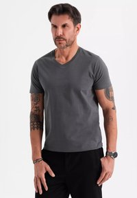 Šedé tričko s véčkovým výstřihem vyrobené z měkkého materiálu, s krátkým rukávem a přiléhavým střihem. Na obrázku jsou tattoované paže a hodinky na zápěstí.