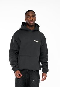 Sudadera con capucha negra, hecha de un material suave, que presenta un bolsillo frontal y la palabra "MULTIPLY" en beige claro. Ajuste casual con puños acanalados.