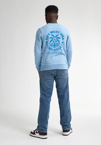 Felpa azzurra con stampa grafica che raffigura una palma e testo sul retro. Abbinata a jeans blu e sneakers nere.