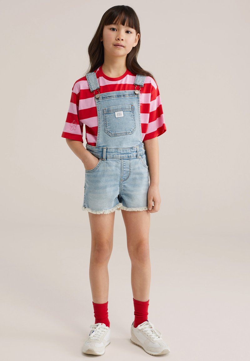 Noor tüdruk seisab käed taskutes, seljas punase ja roosa triibuline särk, hele teksariidest lühikeste overallidega, punased sokid ja valged tossud.