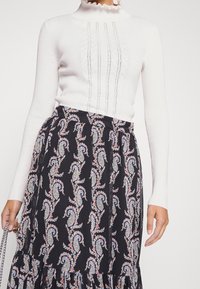 Eine Frau trägt ein weißes, langärmliges Strickoberteil mit Spitzenakzenten und hohem Kragen, kombiniert mit einem schwarzen Rock, der pastellfarbene Paisley-Muster aufweist.