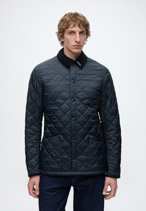 Homme portant une veste matelassée noire avec un col noir, devant boutonné et poches latérales, debout devant un fond blanc uni.